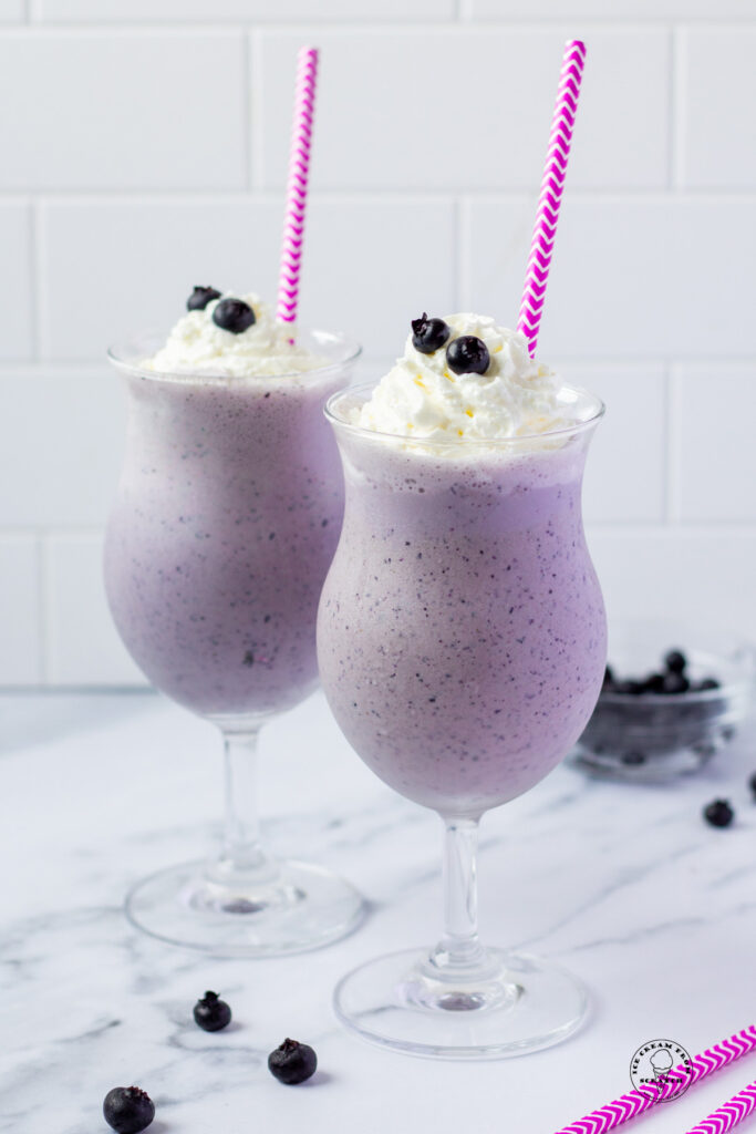 Two blueberry milkshakes in hurricane glasses with purple and white striped straws