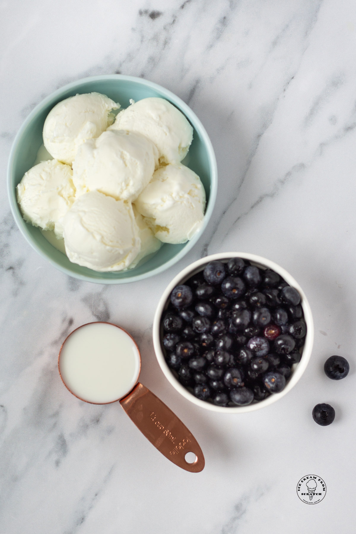 a bowl of scooped vanilla ice cream, a smaller bowl of fresh blueberries, and a measuring cup of milk on a marble countertop