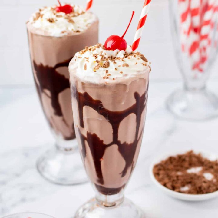Two footed milkshake glasses filled with chocolate milkshake, swirled with chocolate syrup, and topped with whipped cream, cherries, and shaved chocolate