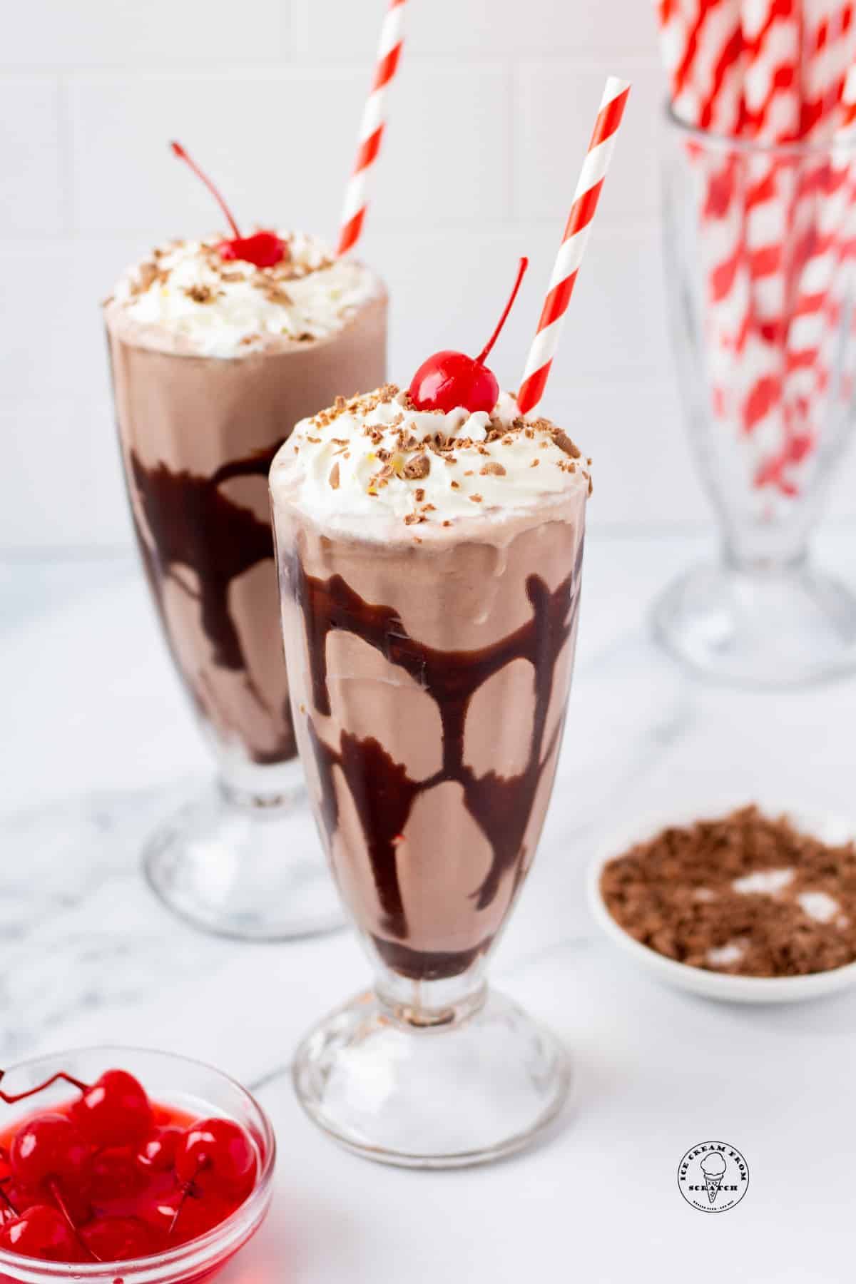 Two footed milkshake glasses filled with chocolate milkshake, swirled with chocolate syrup, and topped with whipped cream, cherries, and shaved chocolate