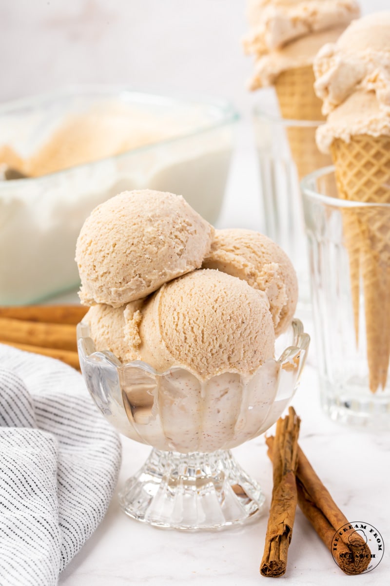 four scoops of cinnamon ice cream in a footed glass dish. Cinnamon sticks are on the counter in front of the dish. Two cones filled with ice cream are in the background.