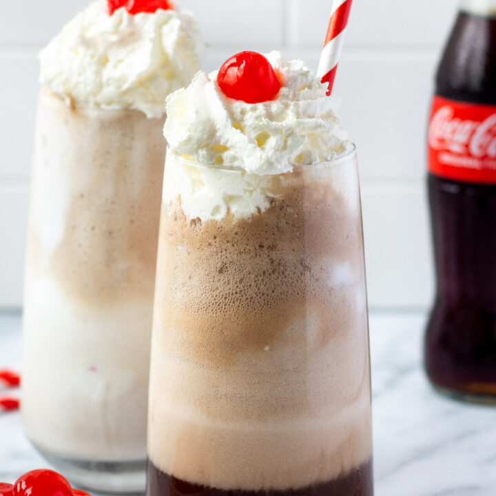 Two tall coke floats topped with whipped cream and a cherry.