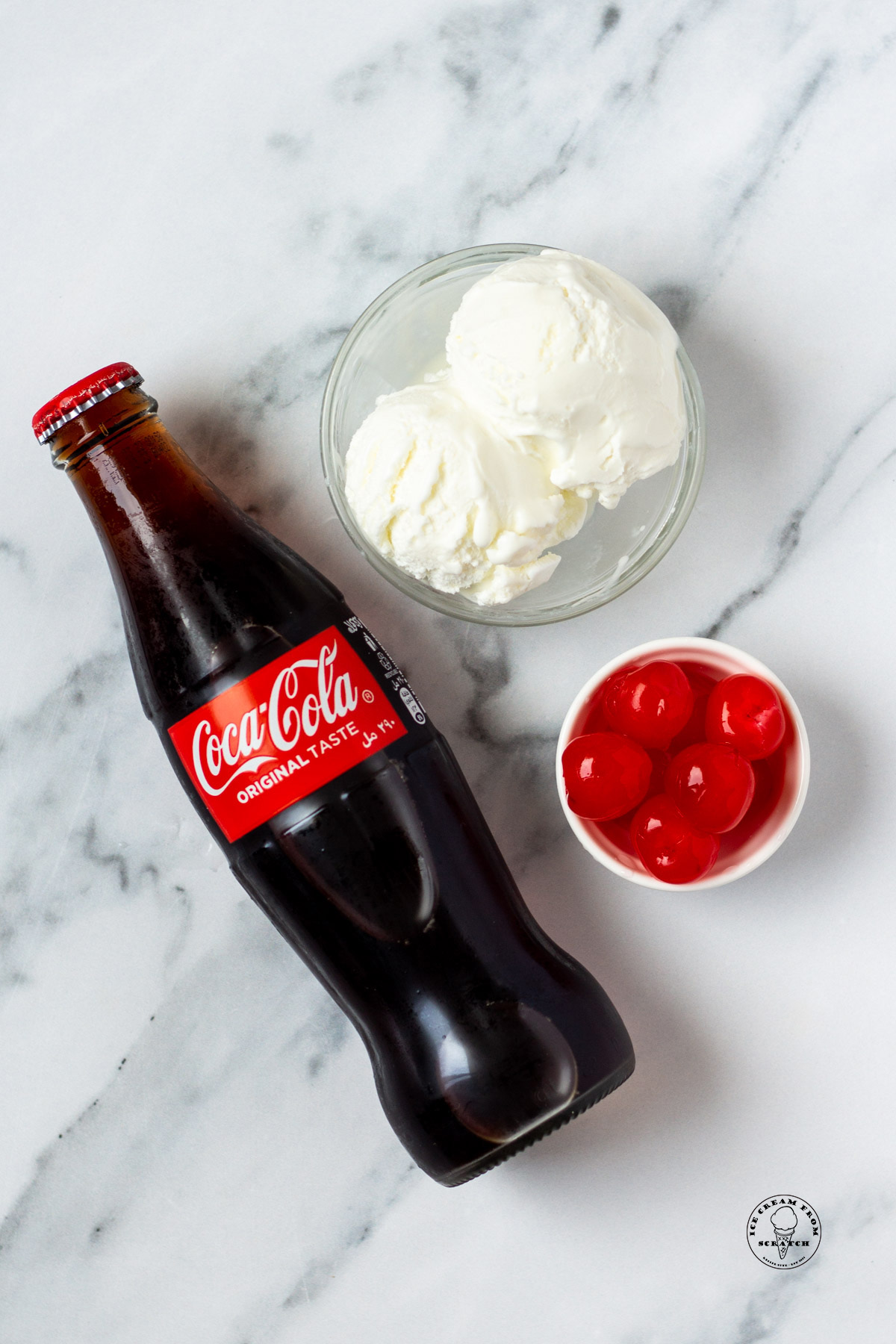 a bowl of vanilla ice cream, a smaller bowl of cherries, and a glass bottle of cocoa cola, viewed from above on a marble counter