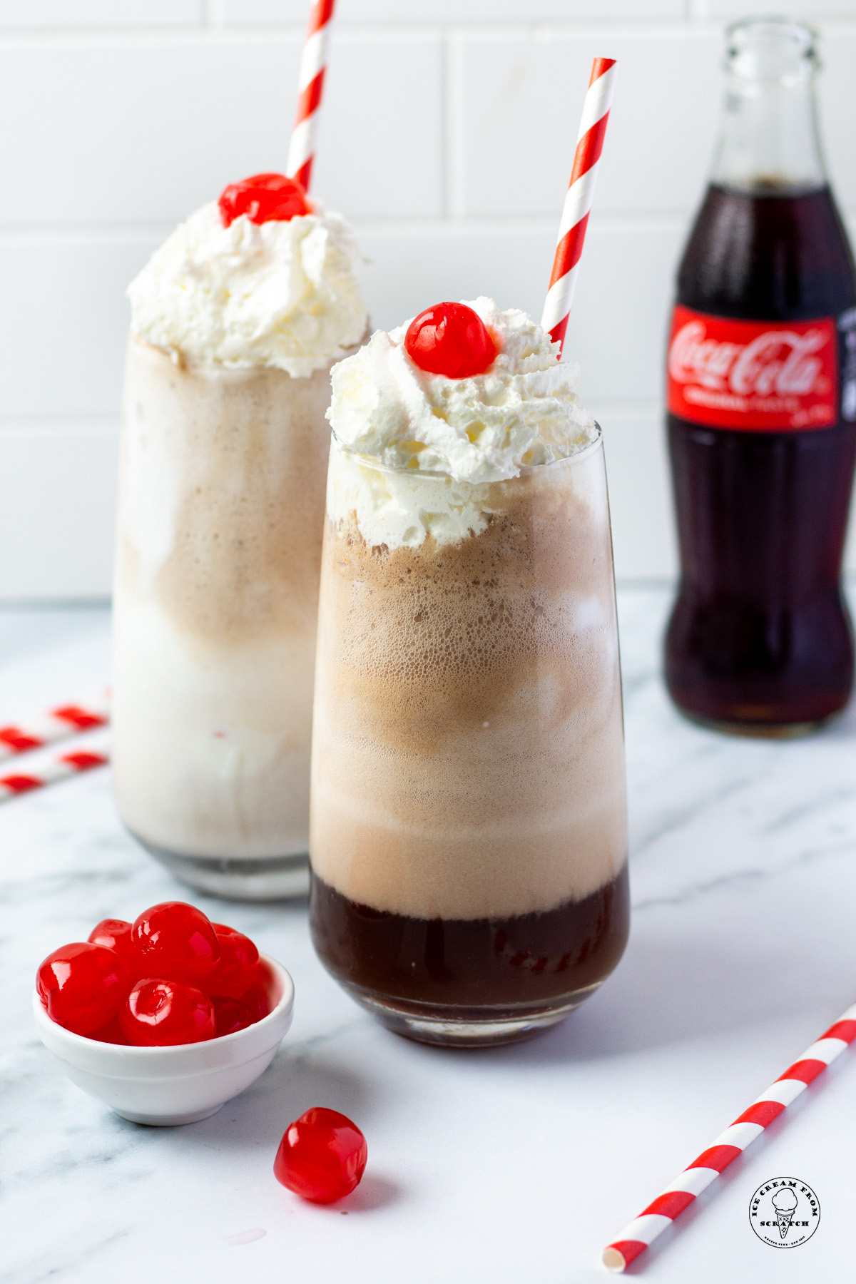 two tall glasses filled with coca cola floats with whipped cream and cherries. Each has a red and white striped straw. A glass bottle of coke is in the background