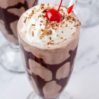 a milkshake glass with a chocoalte syrup drizzle inside, filled with a hersheys milkshake, topped with whipped cream, chocolate shavings, and a cherry.