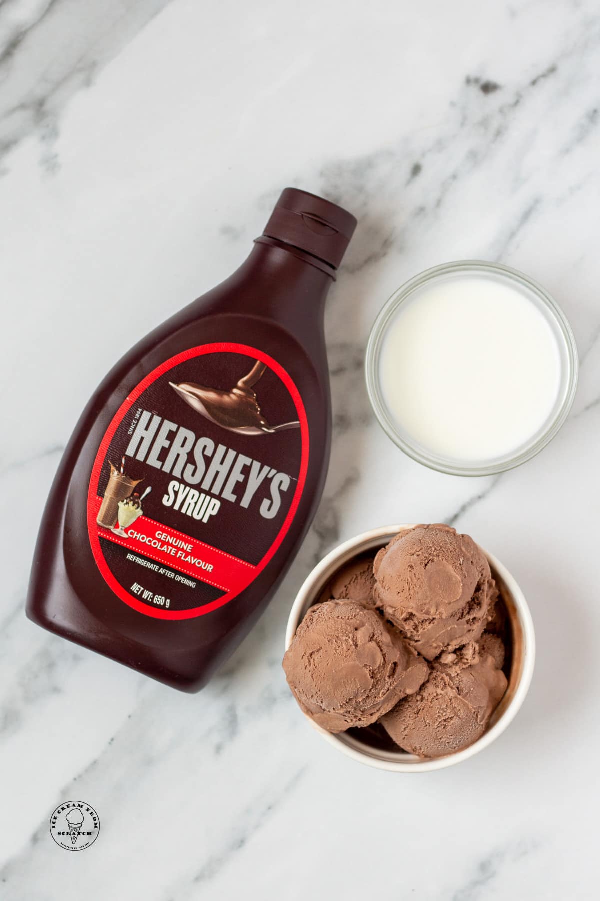 a bottle of hershey's syrup, a bowl of chocolate ice cream, and a bowl of milk, on a marble counter, viewed from above.