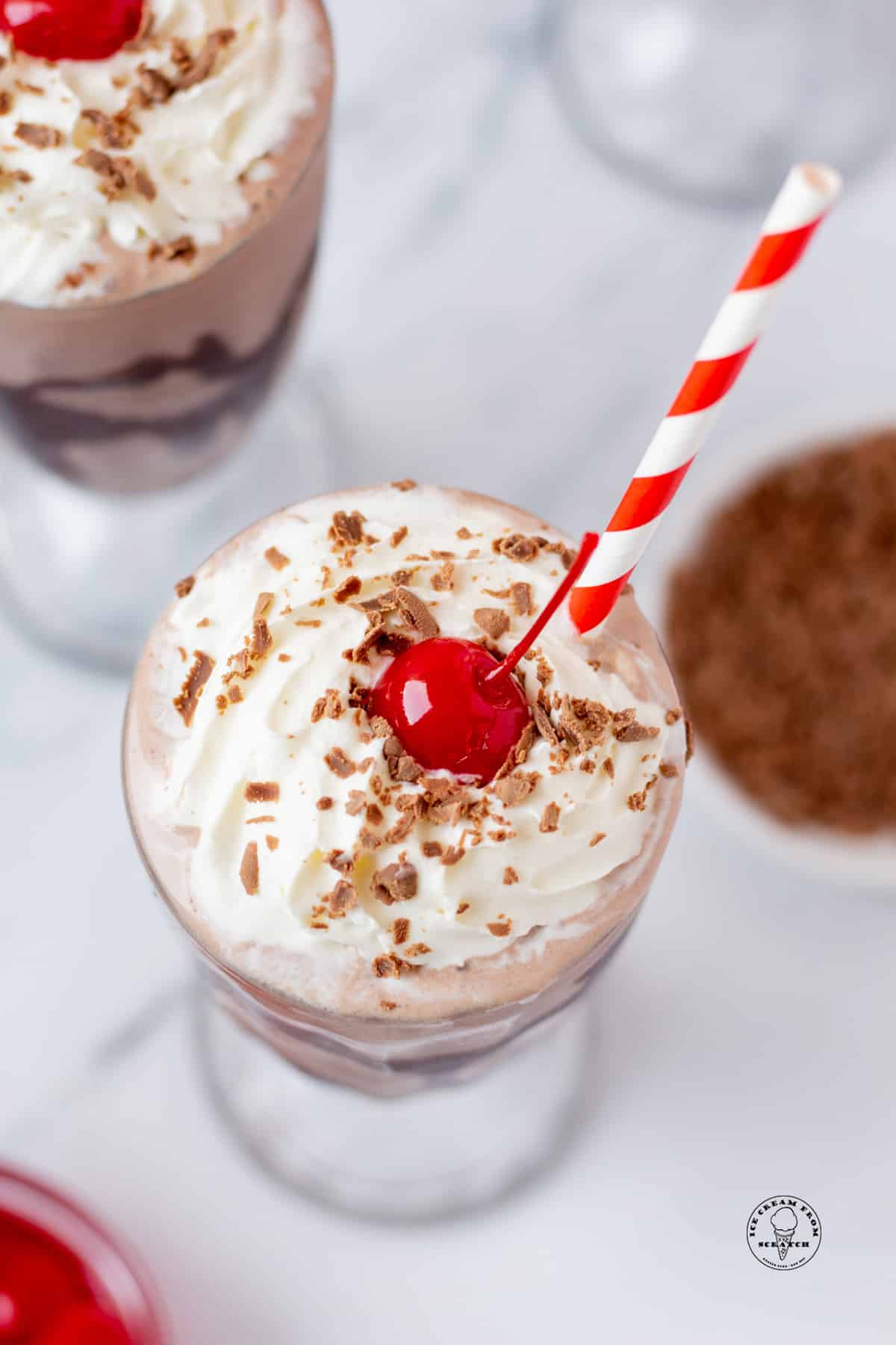 overhead view of a chocolate milkshake topped with whipped cream and a cherry, with a red and white straw.