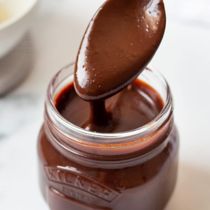 a small glass jar filled with homemade hot fudge. A spoon is lifting some out and the sauce is drizzling back into the jar.