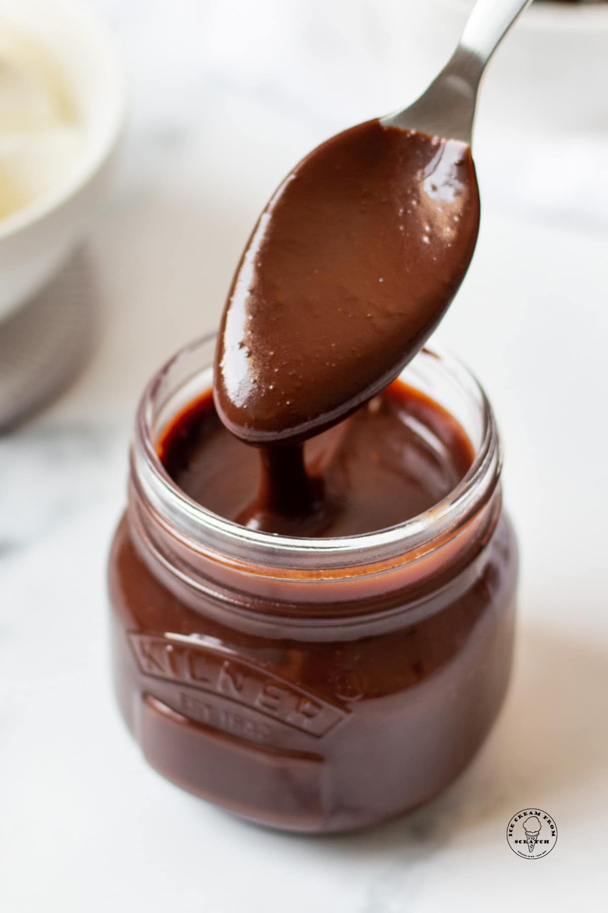 a small glass jar filled with homemade hot fudge. A spoon is lifting some out and the sauce is drizzling back into the jar.