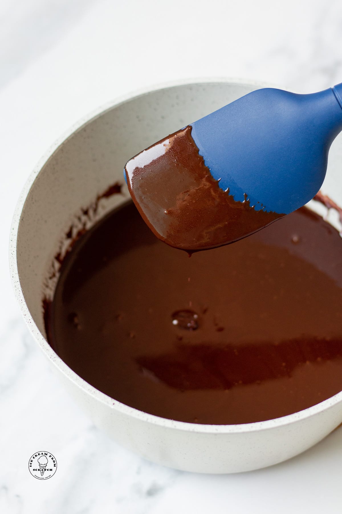 hot fudge sauce in a gray bowl. a blue spatula is holding some up to show the texture and how it sticks to the spoon.