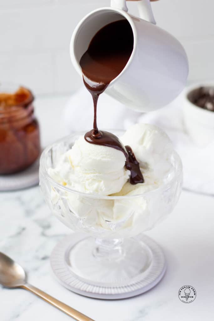 hot fudge in a white pitcher, pouring over a bowl of vanilla ice cream.