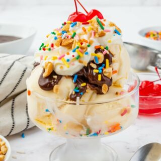 a clear footed ice cream dish filled with a hot fudge sundae topped with peanuts, whipped cream, sprinkles, and two stemmed cherries.