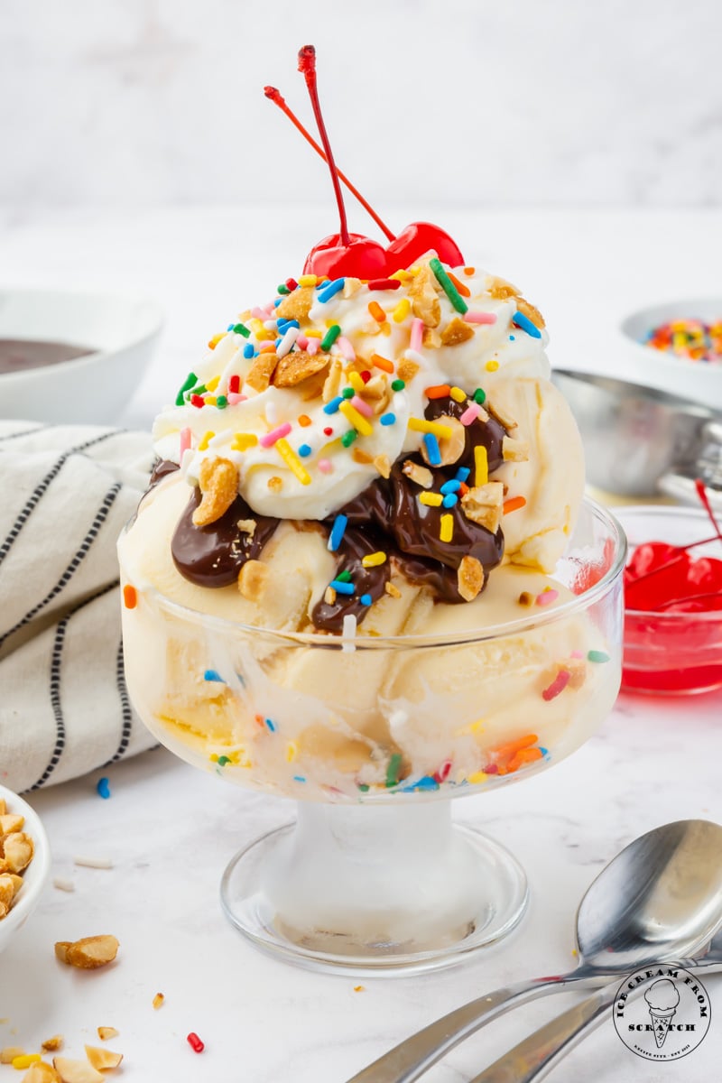 a clear footed ice cream dish filled with a hot fudge sundae topped with peanuts, whipped cream, sprinkles, and two stemmed cherries.