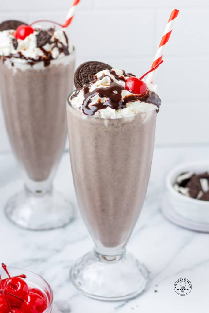 two tall milkshake glasses filled with Oreo milkshakes, topped with whipped cream, chocolate sauce, a cherry, and an oreo. Each has a red and white striped straw