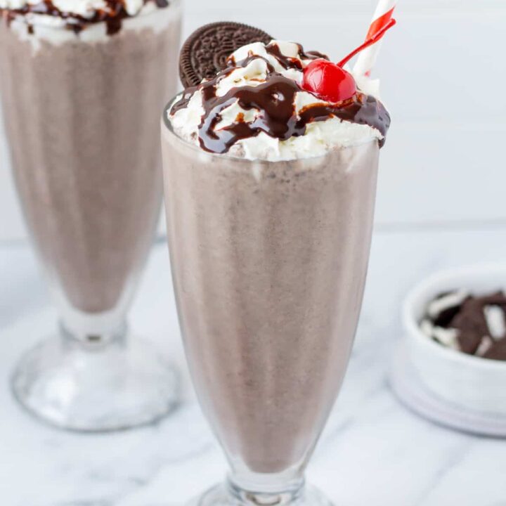 two tall milkshake glasses filled with Oreo milkshakes, topped with whipped cream, chocolate sauce, a cherry, and an oreo. Each has a red and white striped straw