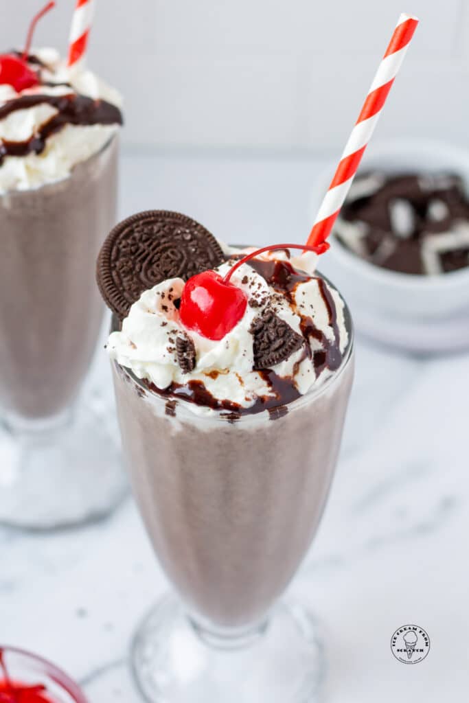 an oreo milkshake in a footed glass, topped with an oreo, oreo crumbs, whipped cream, and cherry.