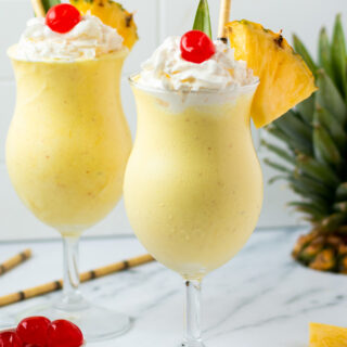 Two pineapple milkshakes in hurricane glasses garnished with a pineapple leaf, whipped cream, cherries, and fresh pineapple wedges.