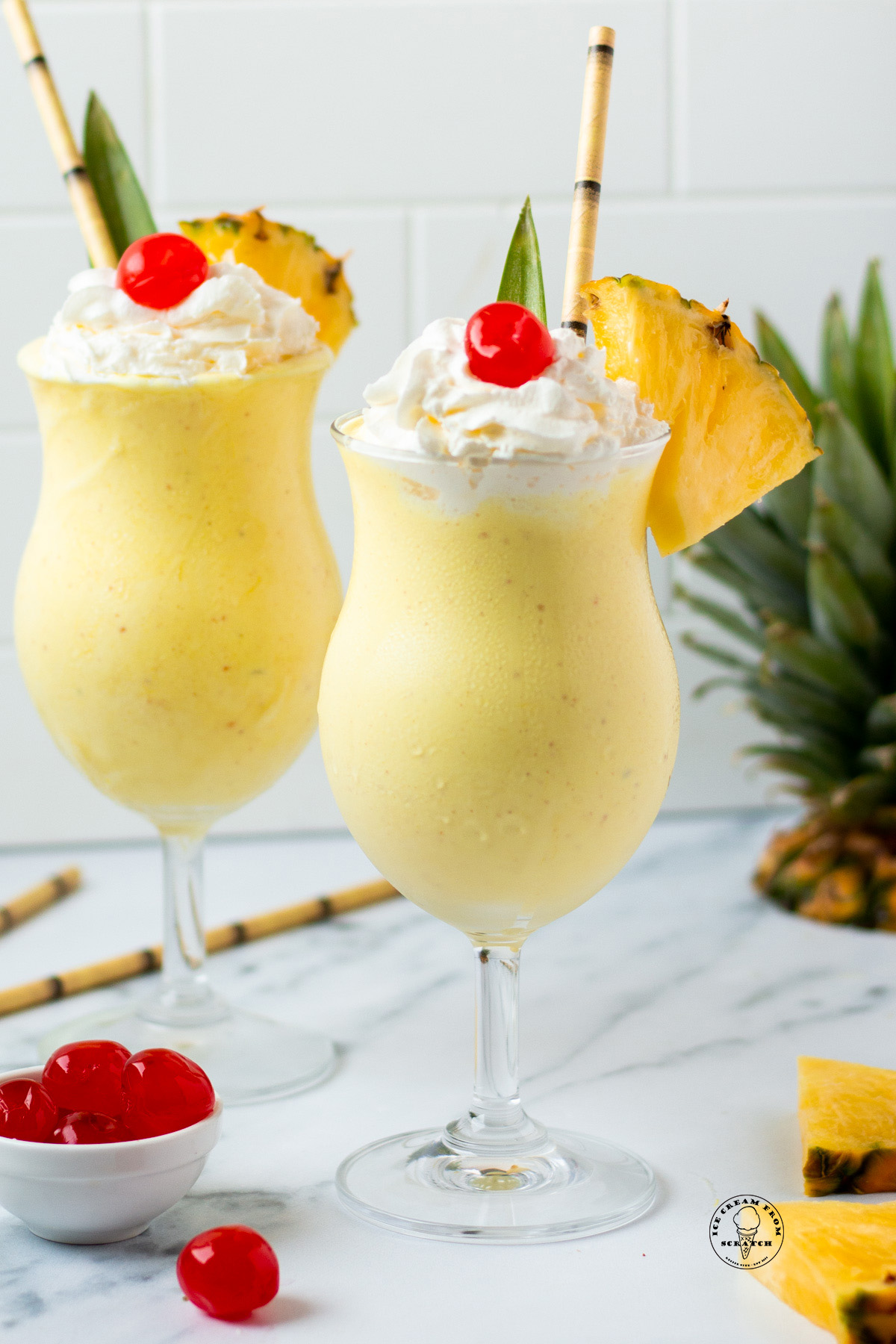 Two pineapple milkshakes in hurricane glasses garnished with a pineapple leaf, whipped cream, cherries, and fresh pineapple wedges.