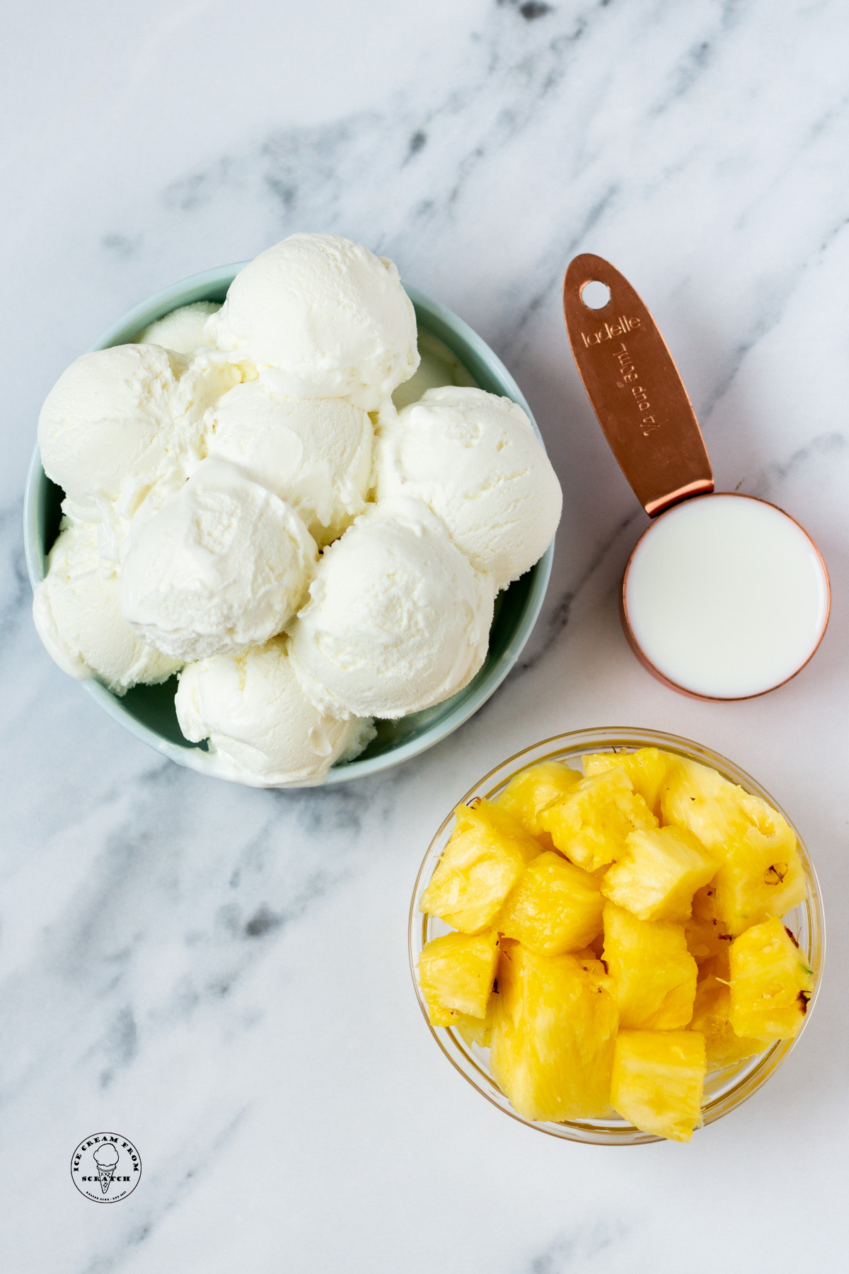 a large bowl of scooped vanilla ice cream, a smaller bowl of diced pineapple, and a measuring cup holding milk, on a marble counter, viewed from above.