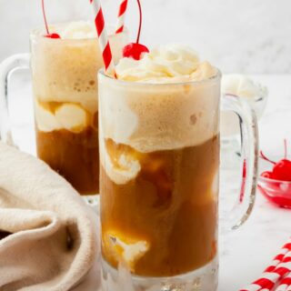 Two frosted rootbeer floats in handled mugs, topped with whipped cream and cherries, with red and white striped straws.