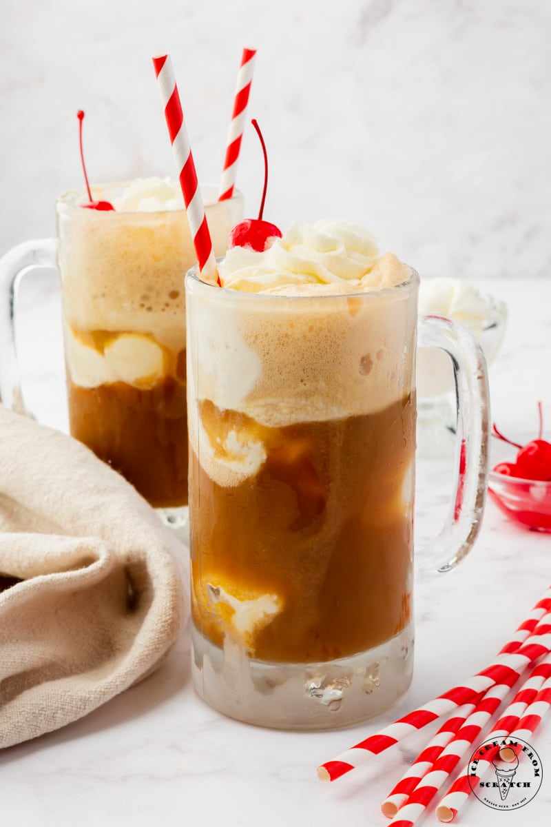 Two frosted rootbeer floats in handled mugs, topped with whipped cream and cherries, with red and white striped straws.