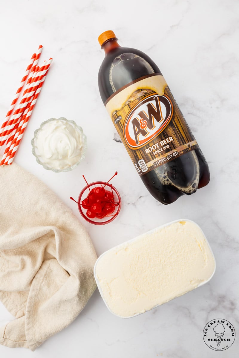 a bottle of a & w root beer, a container of vanilla ice cream, a bowl of stemmed maraschino cherries, and whipped cream on a marble counter, viewed from above.