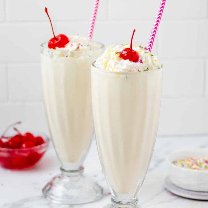 two old fashioned milkshake glasses filled with vanilla milkshake, topped with whipped cream, cherries, and rainbow sprinkles