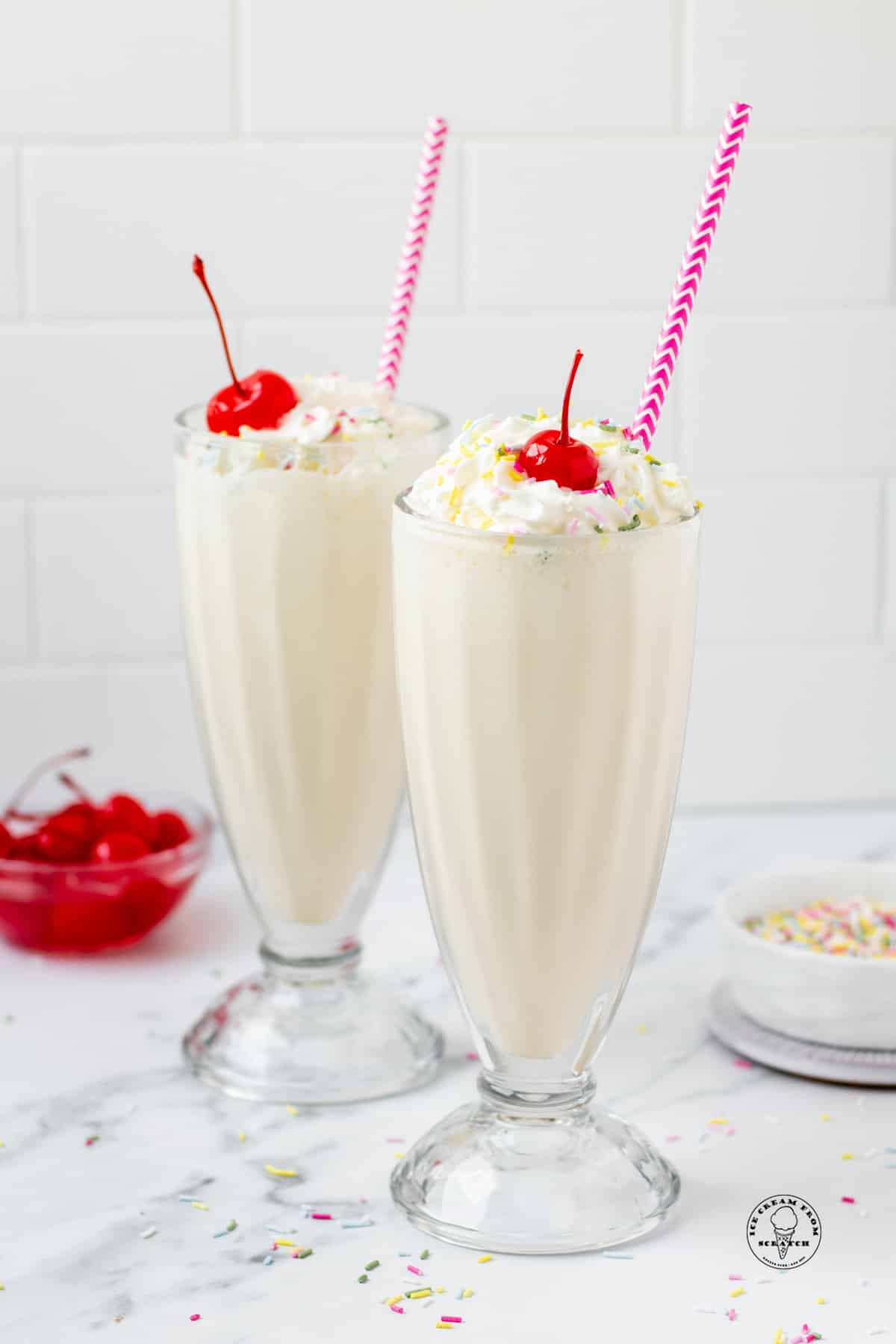 two old fashioned milkshake glasses filled with vanilla milkshake, topped with whipped cream, cherries, and rainbow sprinkles