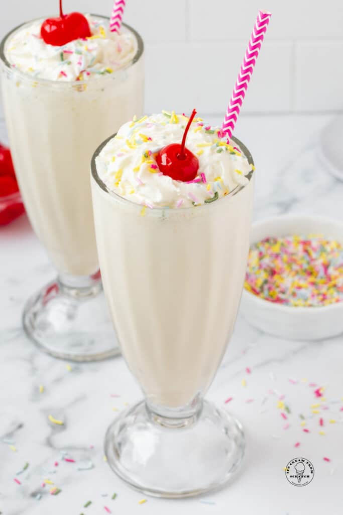 two vanilla milkshakes with purple straws, topped with whipped cream, sprinkles, and cherries