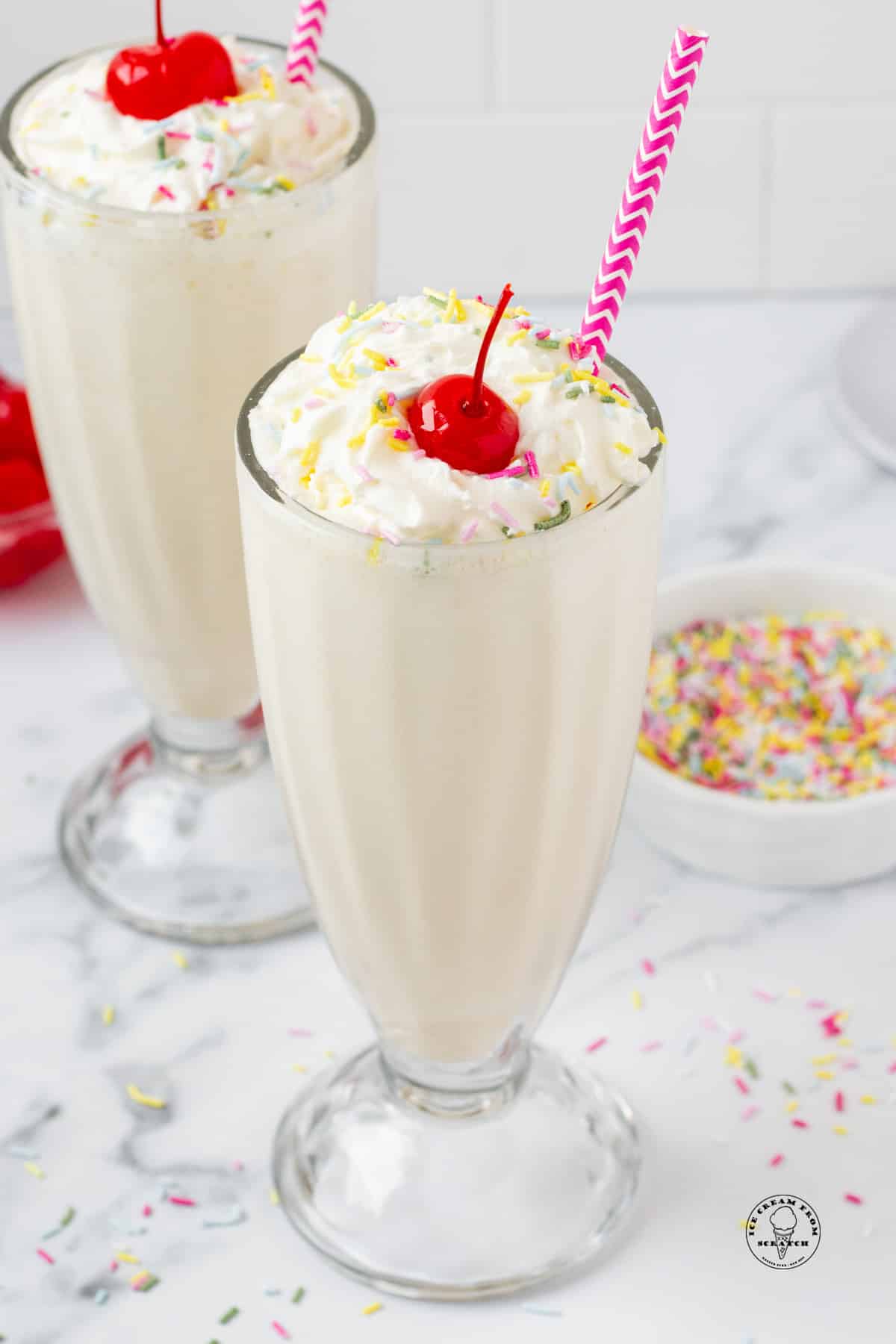 two vanilla milkshakes with purple straws, topped with whipped cream, sprinkles, and cherries
