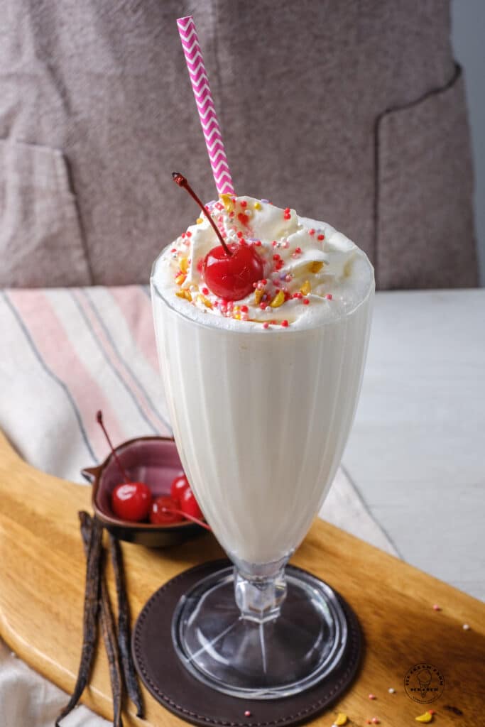 a footed milkshake glass on a leather coaster on a wooden cutten board. The glass is filled with a vegan vanilla milkshake topped with sprinkles, whipped cream, and a cherry.