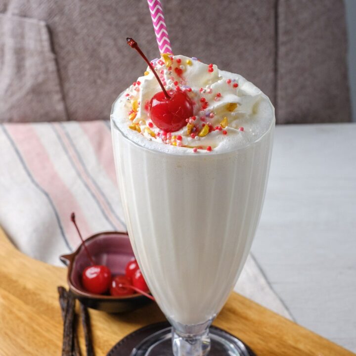 a footed milkshake glass on a leather coaster on a wooden cutten board. The glass is filled with a vegan vanilla milkshake topped with sprinkles, whipped cream, and a cherry.