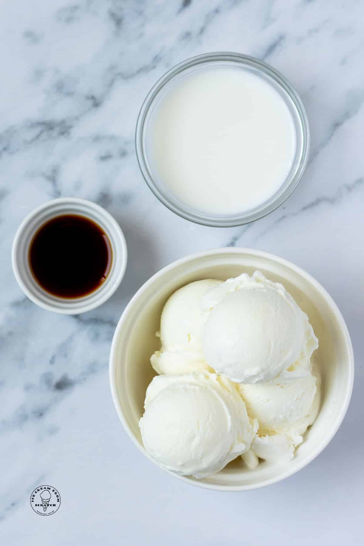 Vegan ice cream, a bowl of vanilla extract, and a bowl of non-dairy milk on a marble counter, viewed from above.