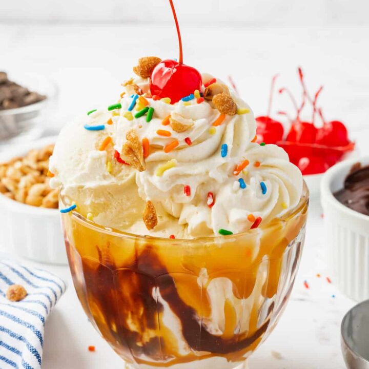 a glass sundae dish filled with a caramel sundae topped with whipped cream, sprinkles, and a cherry.