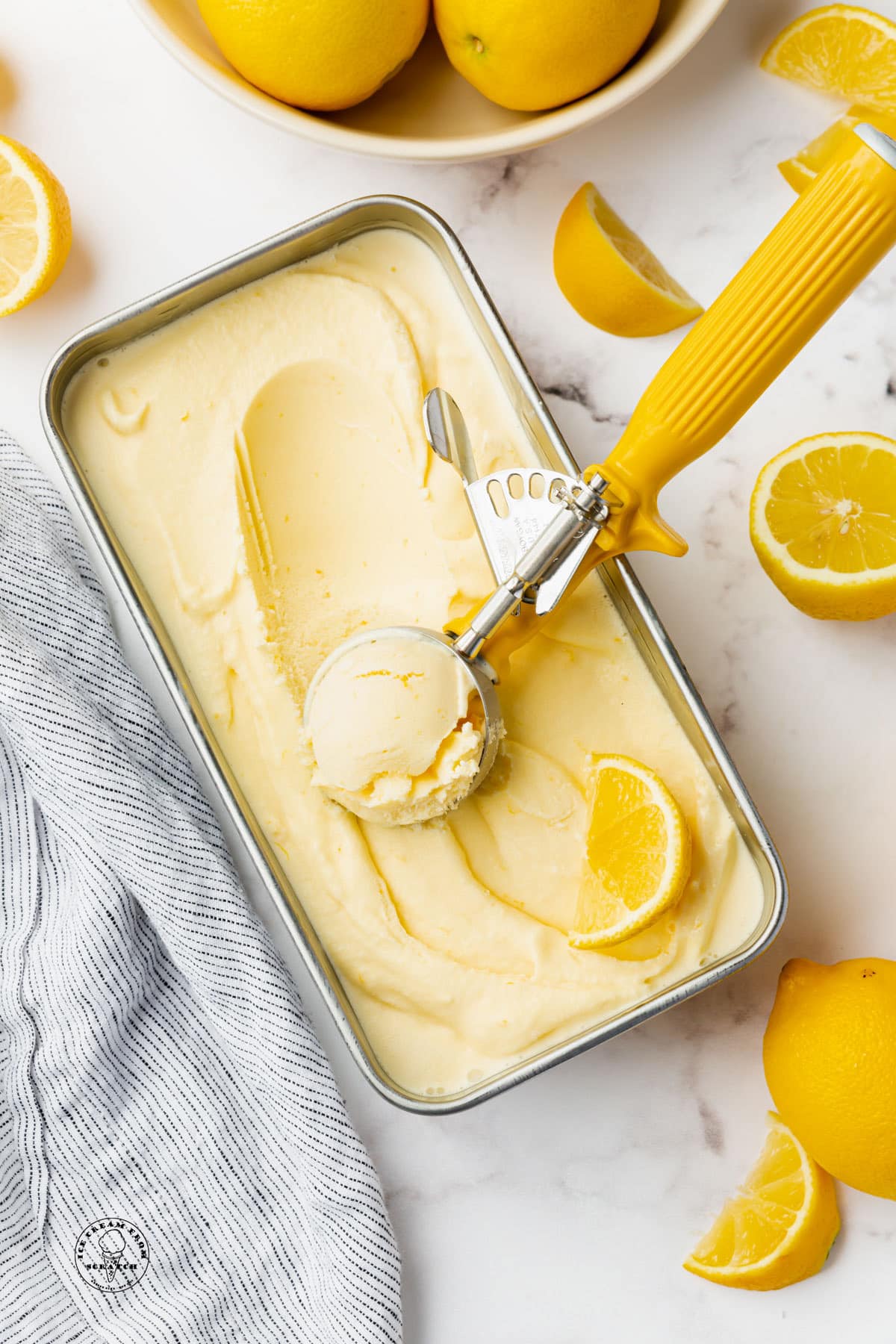 homemade lemon custard ice cream in a metal pan. A yellow-handled ice cream scoop is serving it. Lemon slices are on the counter next to the container.