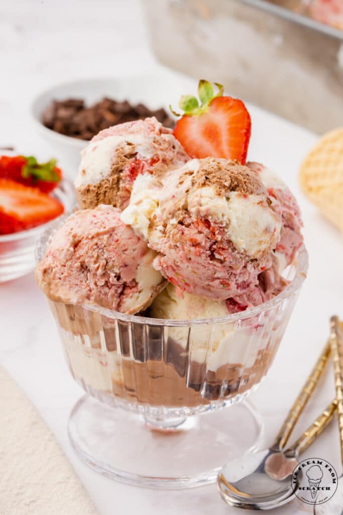 homemade no churn neapolitan ice cream, scooped into a glass, ridged, footed ice cream dish. Two spoons are on the table next to the glass. Dishes of sliced strawberries and chocolate chips are in the background