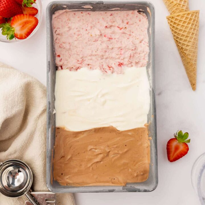 Strawberry, vanilla, and chocolate ice cream, layered into a metal loaf pan, viewed from above. Next to the pan of ice cream are two sugar cones, a bowl of strawberries, a bowl of chocolate chunks, and a red and silver ice cream scoop.