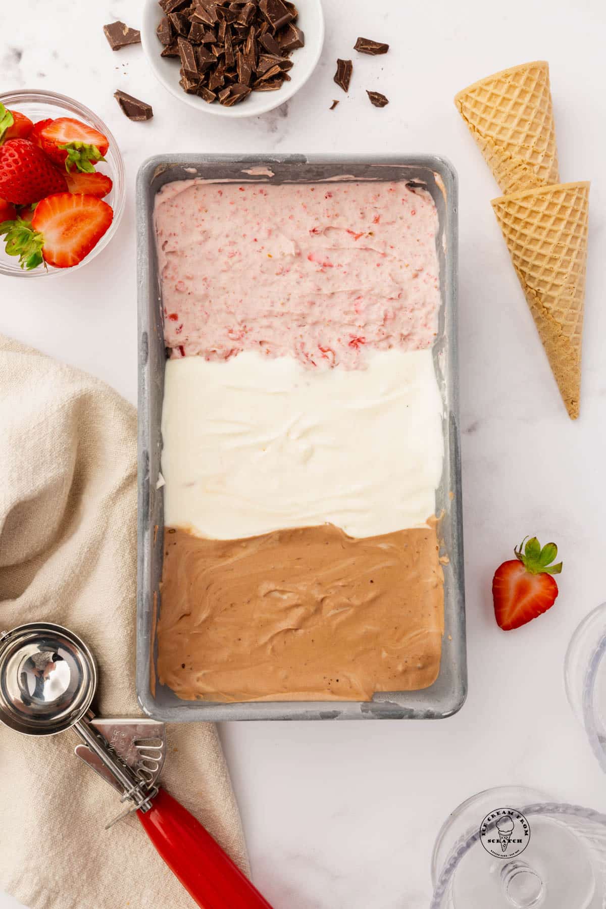 Strawberry, vanilla, and chocolate ice cream, layered into a metal loaf pan, viewed from above. Next to the pan of ice cream are two sugar cones, a bowl of strawberries, a bowl of chocolate chunks, and a red and silver ice cream scoop.