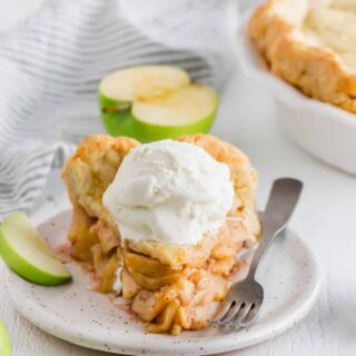 a thick slice of homemade apple pie with a scoop of vanilla ice cream on top. A fork and a slice of green apple are on the plate.