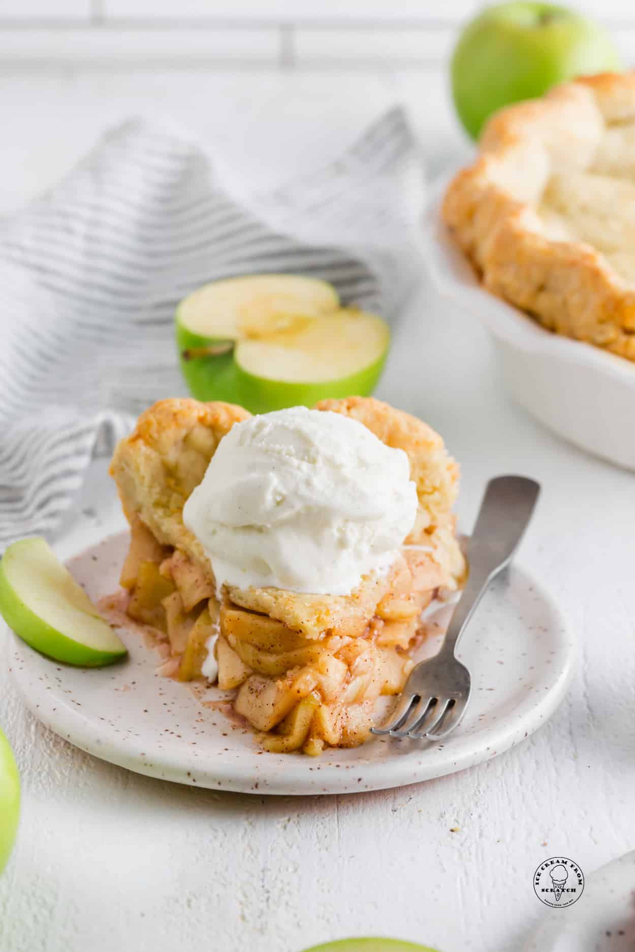 a thick slice of homemade apple pie with a scoop of vanilla ice cream on top. A fork and a slice of green apple are on the plate.