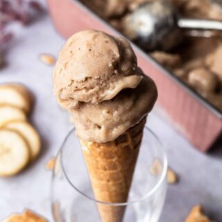homemade banana ice cream scooped into a sugar cone and propped up in a blass. in the background is the pan of ice cream, banana slices, and pieces of cones.