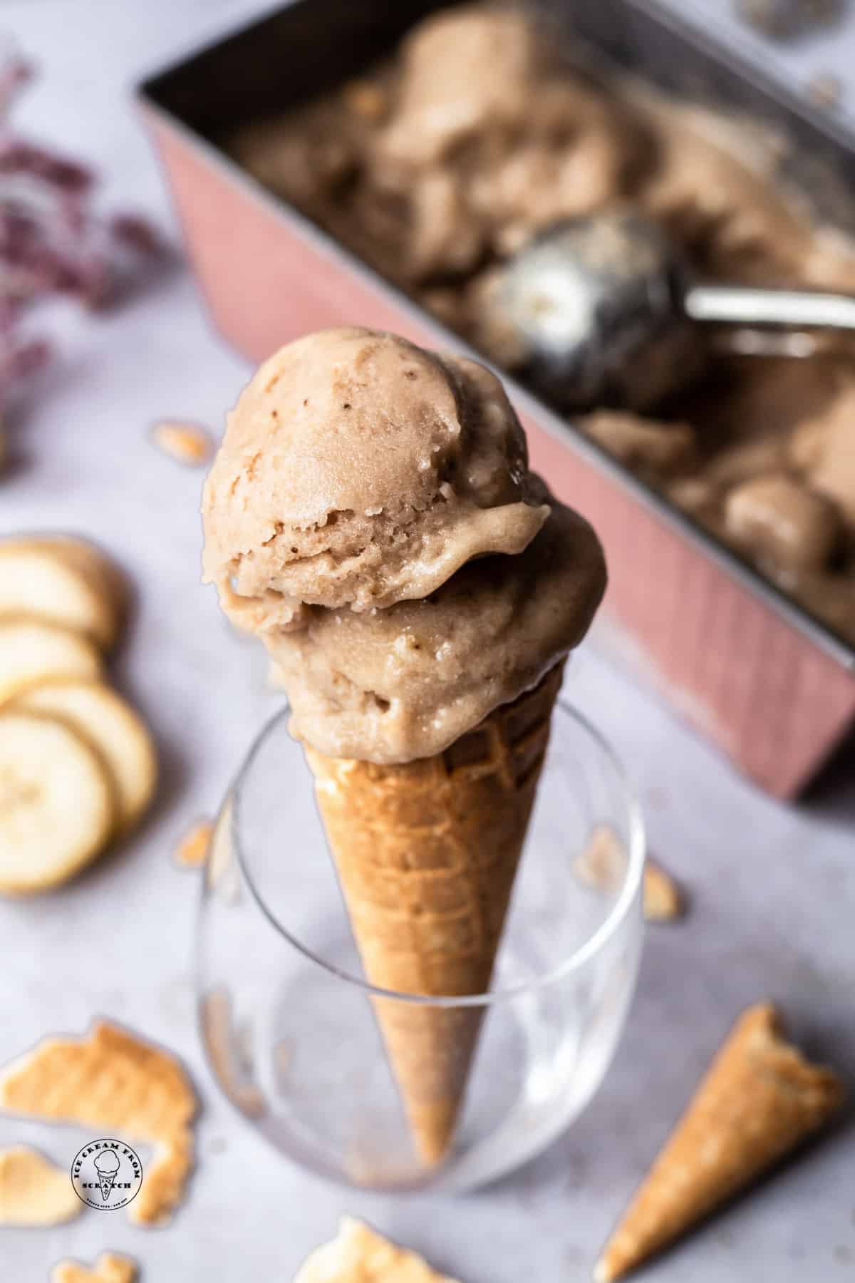 homemade banana ice cream scooped into a sugar cone and propped up in a blass. in the background is the pan of ice cream, banana slices, and pieces of cones.