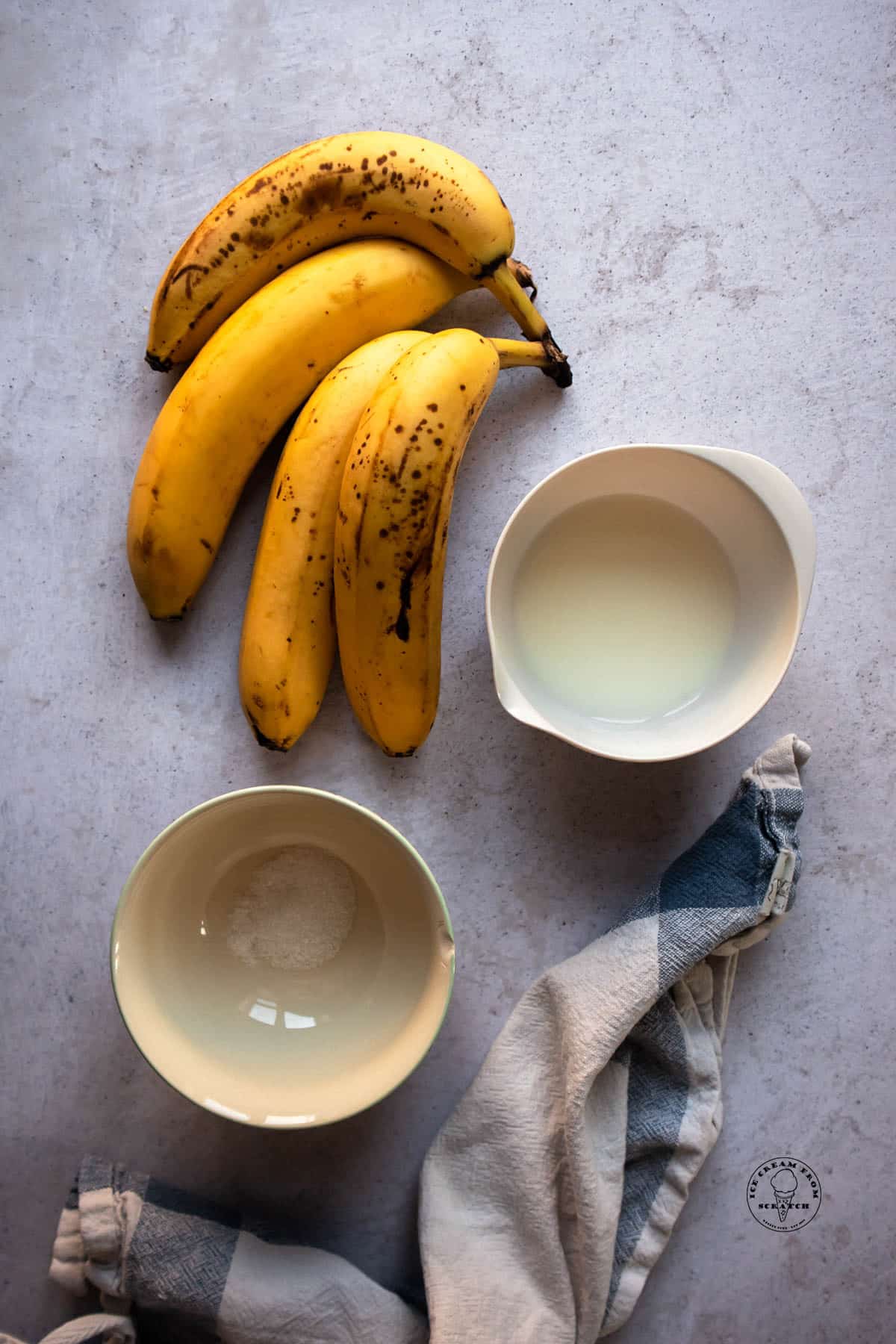 4 ripe bananas, a small bowl of milk, and a small bowl with a pinch of salt, on a concrete counter. A blue plaid dish towel is there as well.