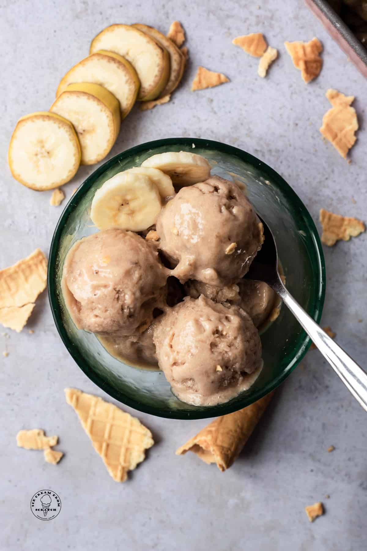 Scoops of banana ice cream (aka nice cream) in a glass bowl, viewed from overhead. Around the bowl are sliced bananas and pieces of ice cream cones.