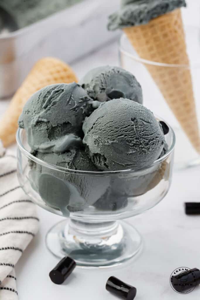 a footed glass bowl filled with scoops of gray ice cream. chunks of black licorice are on the counter next to the dish.