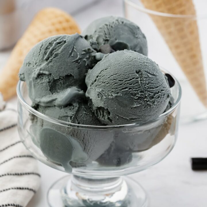 a footed glass bowl filled with scoops of gray ice cream. chunks of black licorice are on the counter next to the dish.
