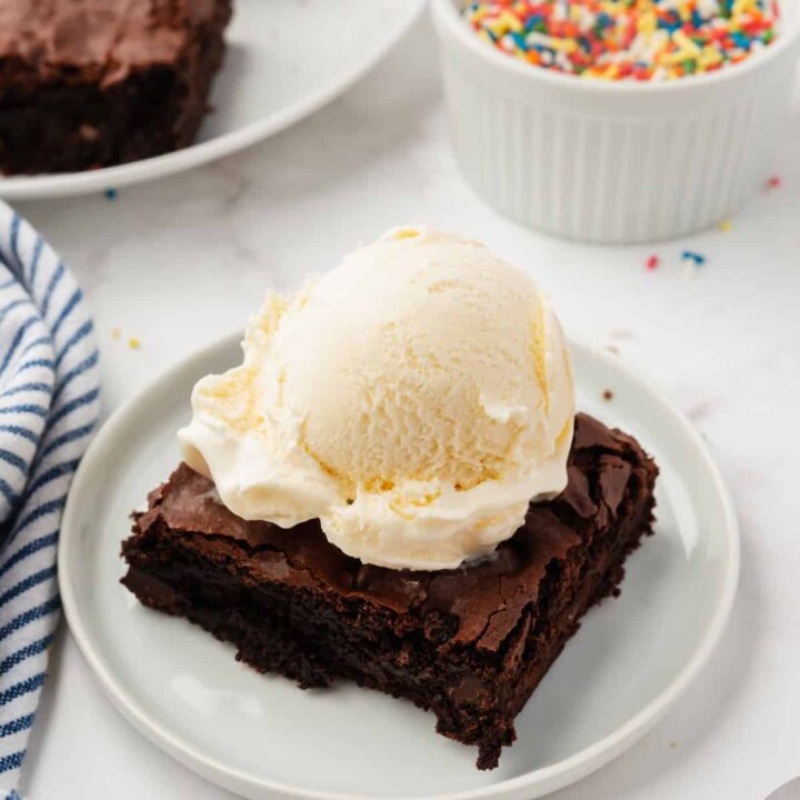 a chocolate brownie on a small plate with a scoop of vanilla ice cream on top. In the background is a bowl of sprinkles.