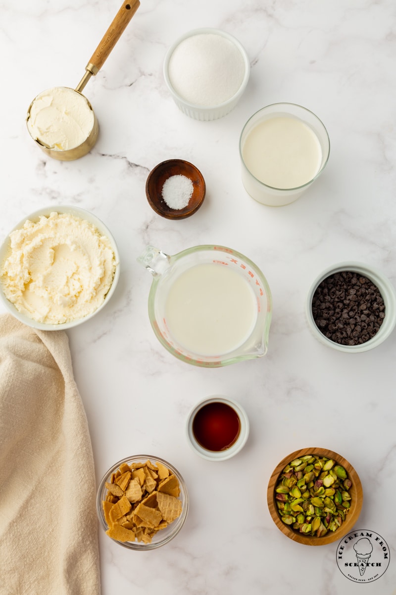 The ingredients needed to make cannoli ice cream, including mascarpone cheese, pistachios, waffle cone pieces, and ricotta cheese. Each ingredient is in a small bowl, arranged on the counter and viewed from overhead.