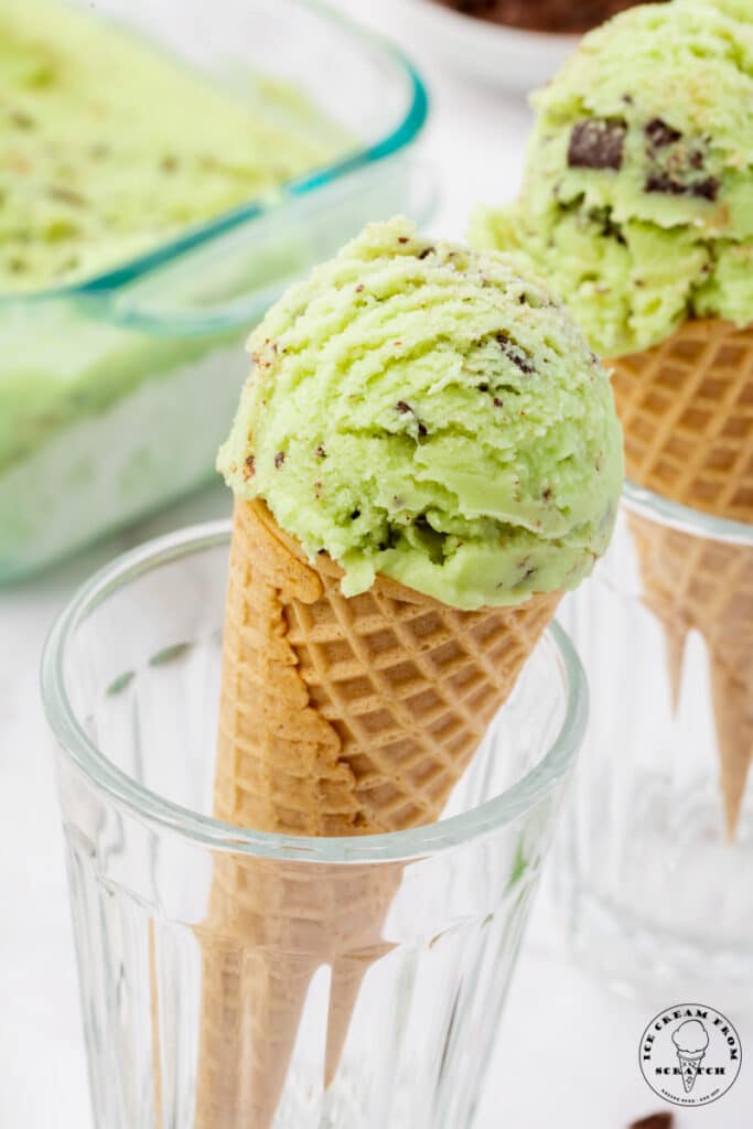 coconut mint ice cream scooped into two sugar cones that are propped up in glasses so that they are upright. The ice cream is green with chocolate chunks in it.