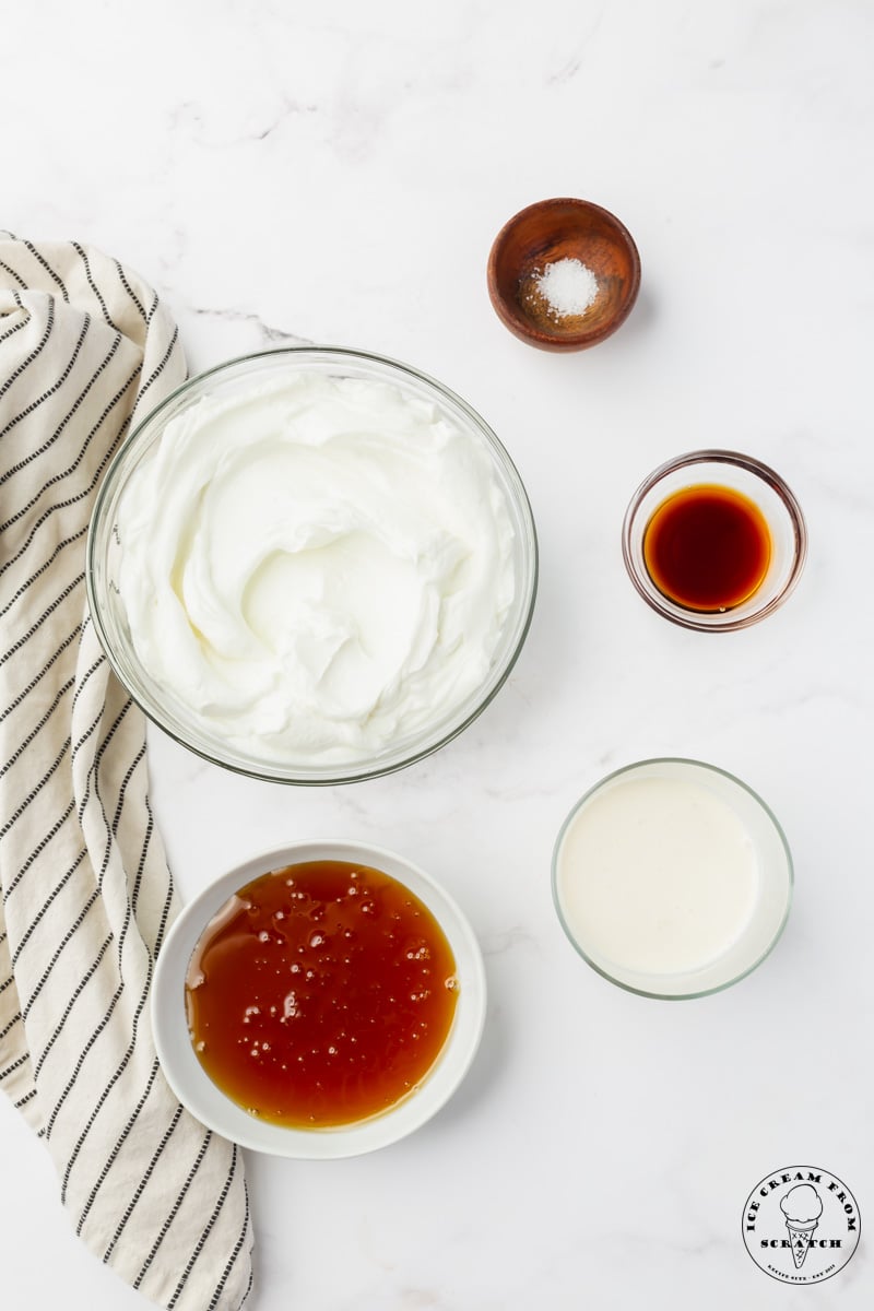 a bowl of green yogurt, a smaller bowl of honey, and three even smaller bowls holding salt, vanilla, and heavy cream. The bowls are arranged on a marble counter, viewed from overhead.